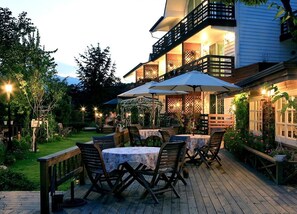 Un patio al aire libre con mesas y sillas, rodeado por una terraza de madera y abundante follaje.