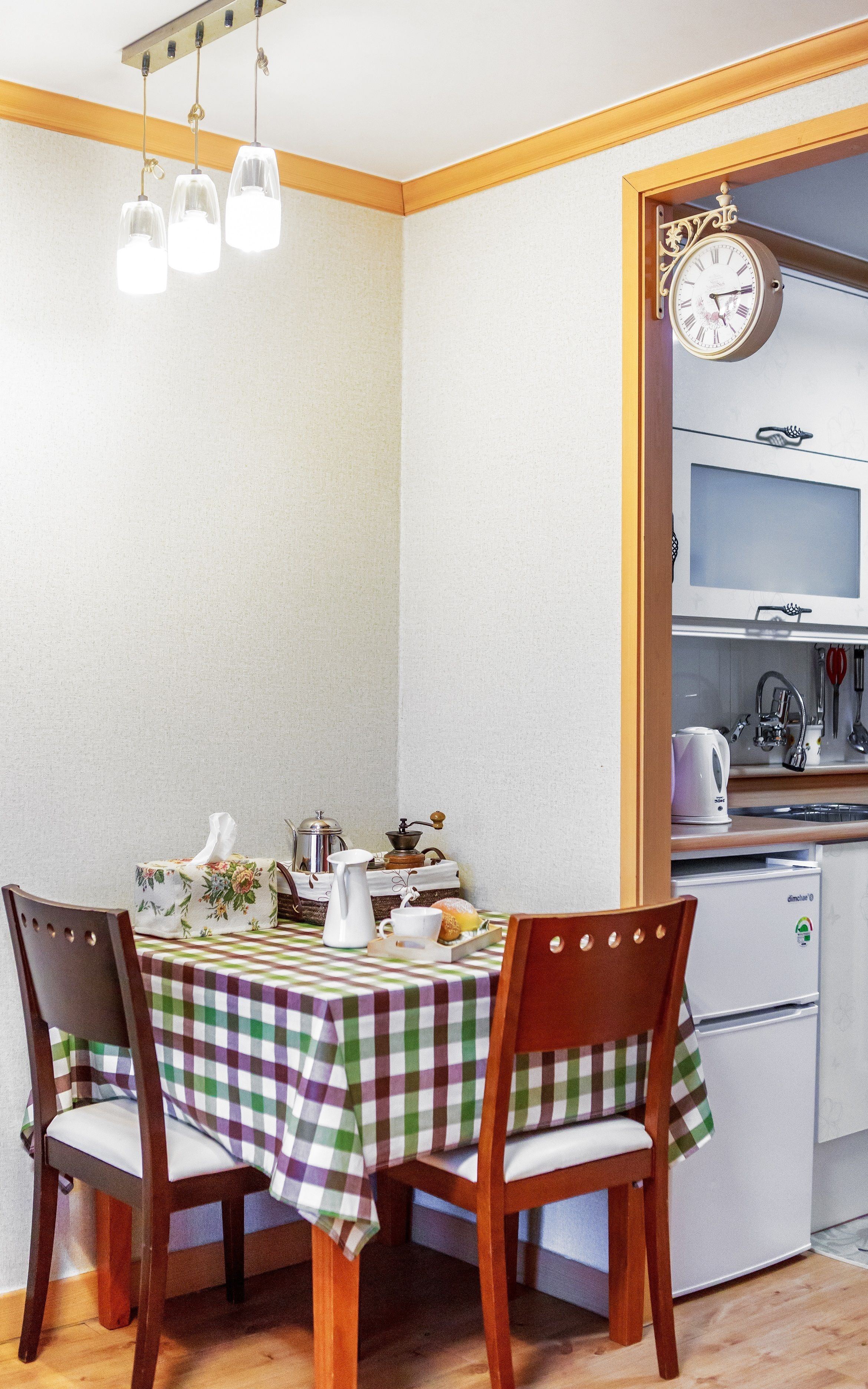 A small kitchen with a table set for two, a clock on the wall, and a refrigerator.
