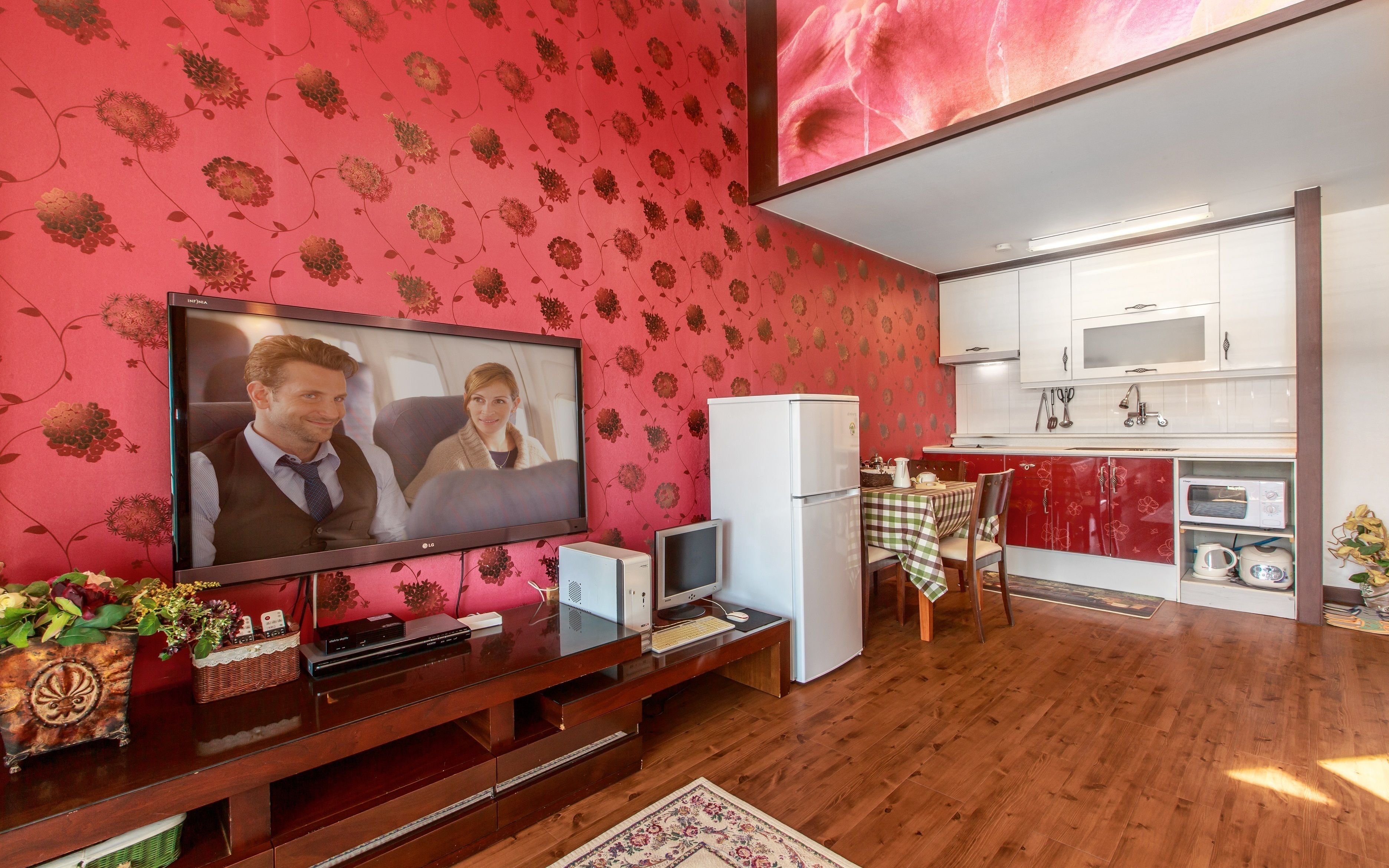 A compact living space with a television, a small refrigerator, a dining area with a table and chairs, and a kitchen with white cabinets and a microwave.