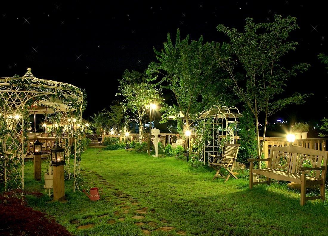 Un jardín bien iluminado con un pabellón, bancos de madera y un camino.