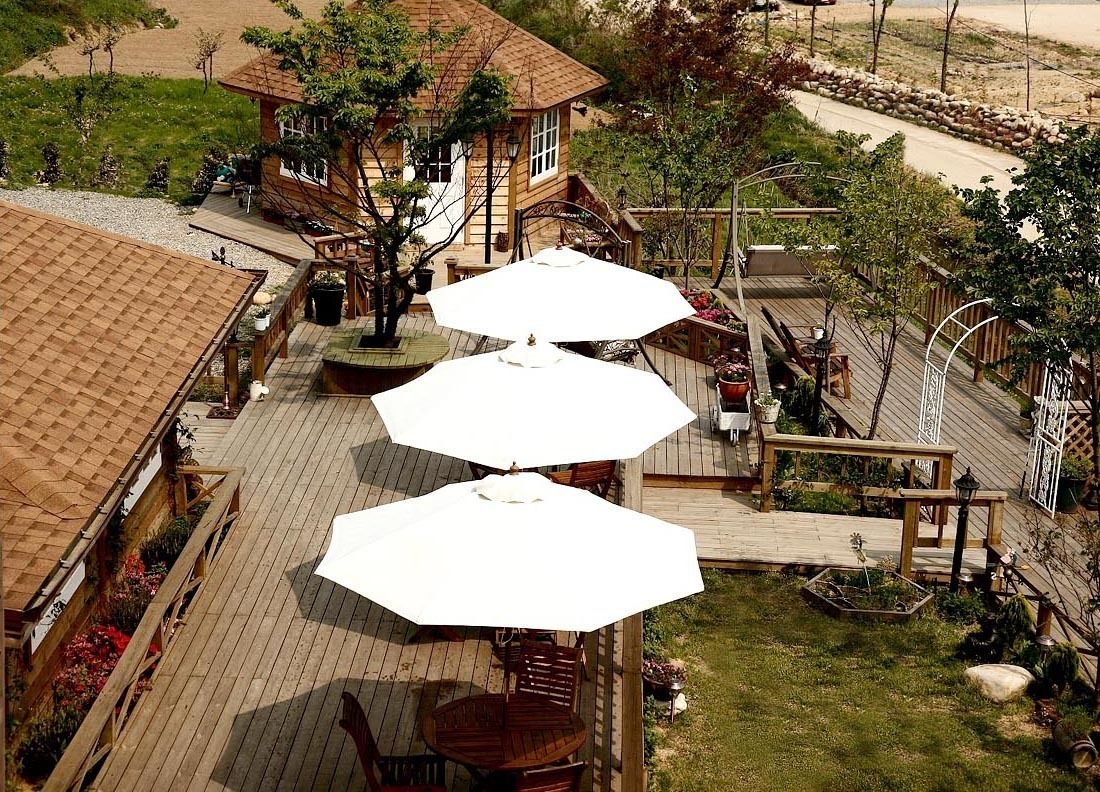 A wooden deck with outdoor seating and umbrellas, surrounded by a landscaped garden and a building with a sloped roof.