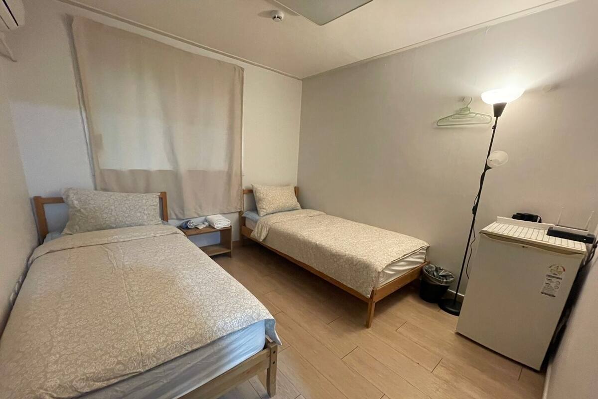 A small, clean room with two single beds, a nightstand, a desk, a lamp, and a refrigerator.