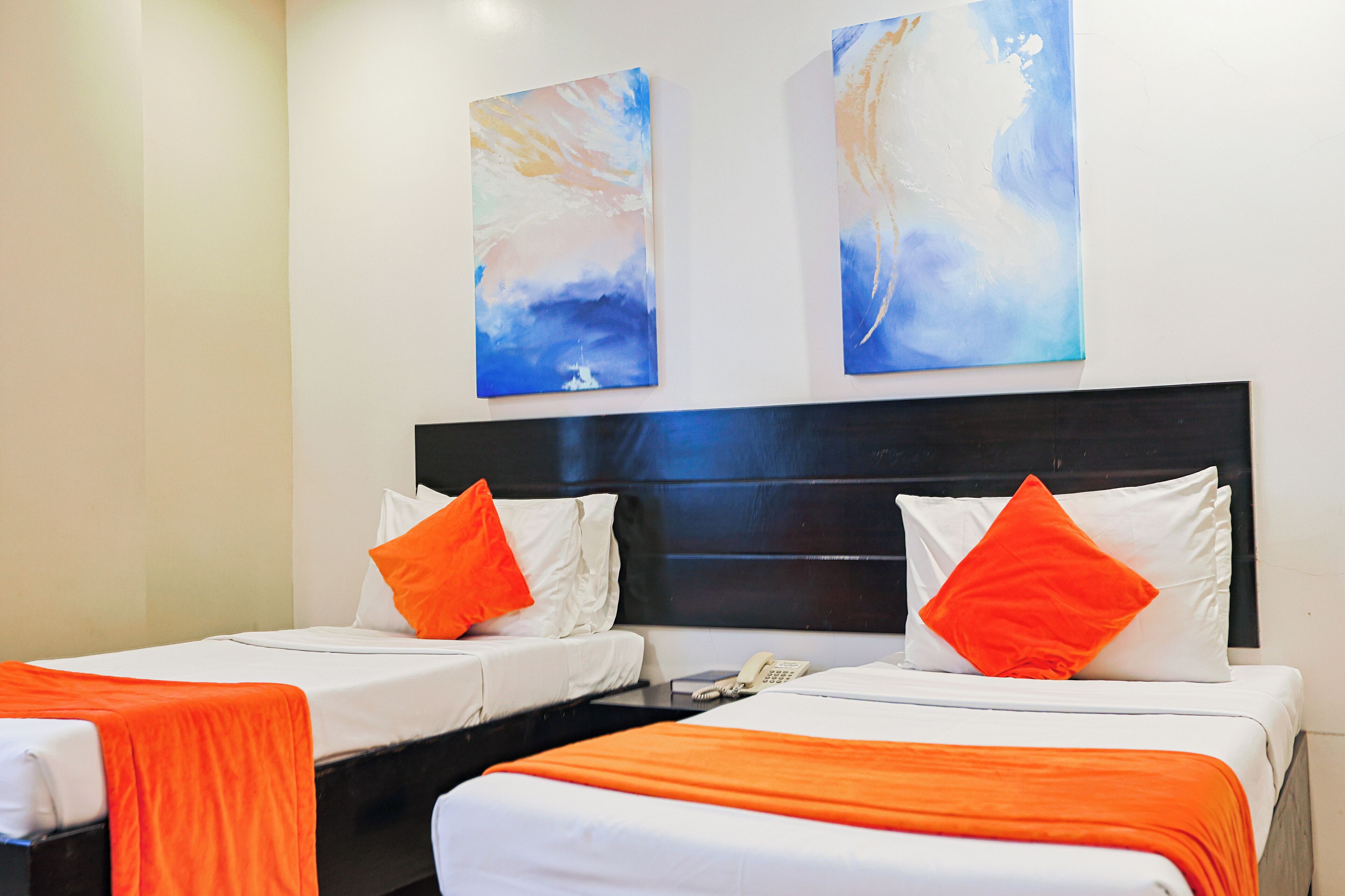 Une chambre d’hôtel avec deux lits, chacun agrémenté de draps blancs et de touches de couleur orange, d’une tête de lit noire et de tableaux abstraits accrochés au mur.