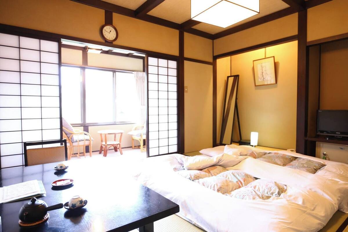 Een traditionele Japanse kamer met tatami vloeren, een bed met wit beddengoed, een televisie en een schuifdeur naar een balkon.