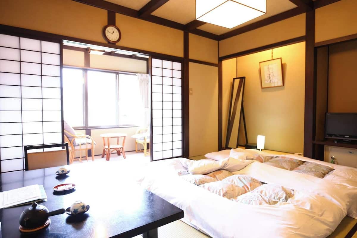 Una habitación tradicional japonesa con suelo de tatami, una cama con ropa blanca, un televisor y un puerta corredera que da a un balcón.