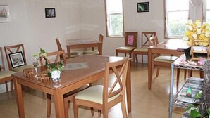A dining area with wooden tables and chairs, framed pictures on the walls, and a window with a view of greenery. - Pension Souvenir (Iki)