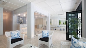 Um hall espaçoso com móveis de vime branco, piso xadrez e vista para um ambiente bem iluminado também com piso xadrez.