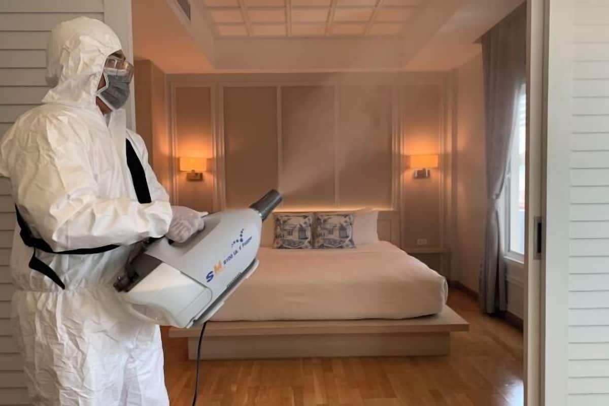 Une personne vêtue d’une combinaison de protection blanche utilise un appareil dans une chambre à coucher équipée d’un lit, de tables de chevet et de lampes fixées au mur.