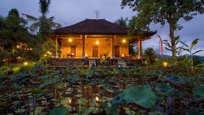A traditional building with a tiled roof, surrounded by a pond with lotus plants, and a few trees in the background. - Subak Tabola Villa (Sidemen)