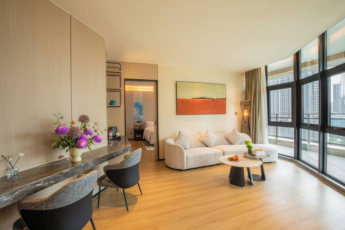 A modern living room with a sofa, chairs, a coffee table, and a large window offering a city view.