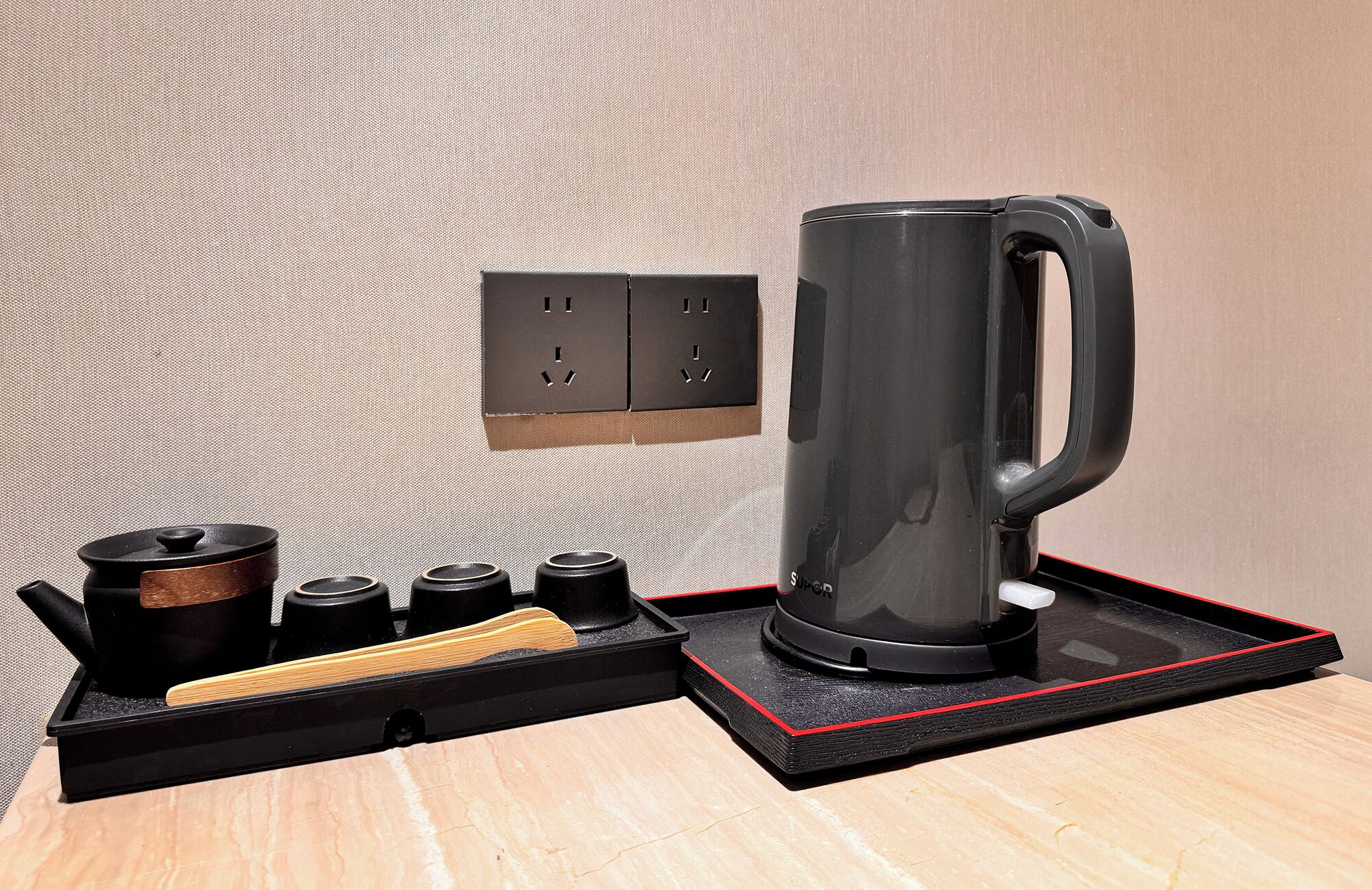Una cafetera, un hervidor y una tetera sobre una bandeja con borde rojo, sobre una mesa de madera frente a una pared con textura.