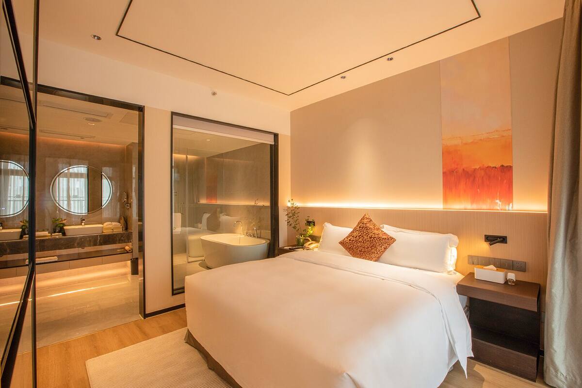 A modern hotel room with a large bed, a glass-enclosed shower area, a bedside table with a lamp, and a wall-mounted artwork.