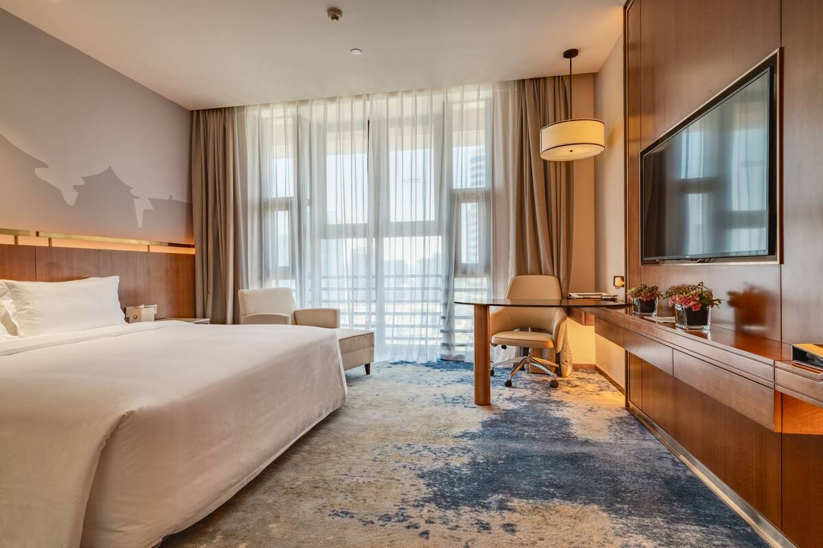 A hotel room with a large bed, a desk with a chair, a TV, and a view of the city through sheer curtains.