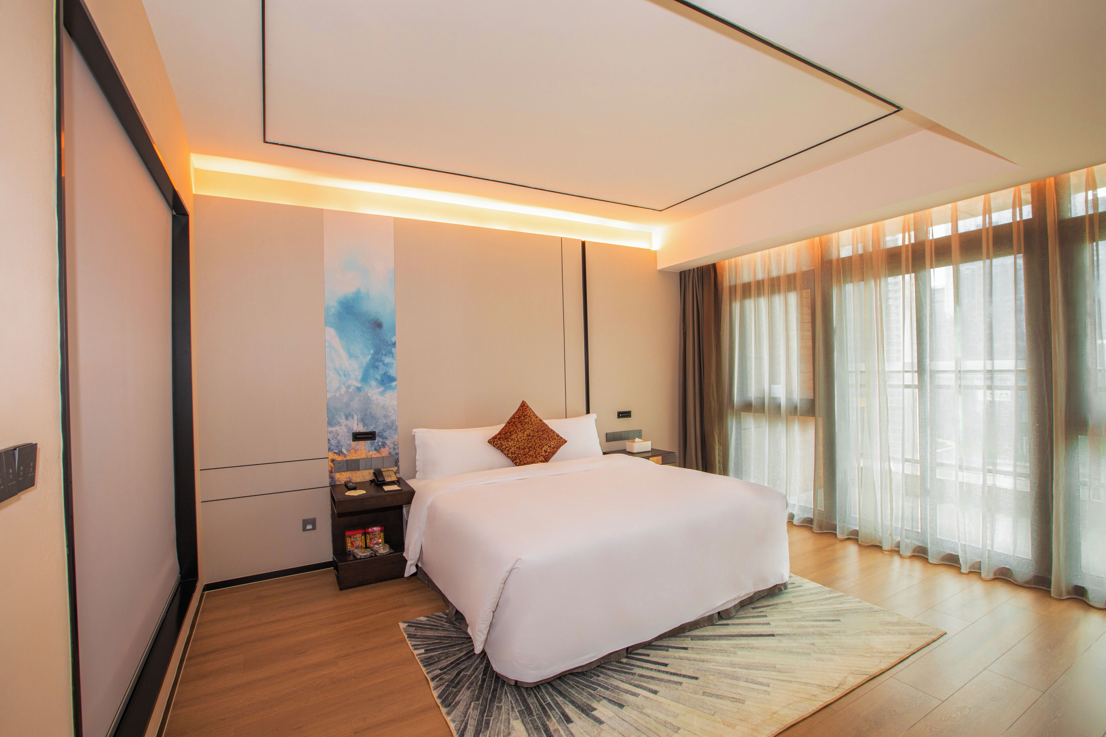 Una habitación de hotel moderna con una cama grande, una mesita de noche, una lámpara de pared y vistas al exterior a través de cortinas traslúcidas.