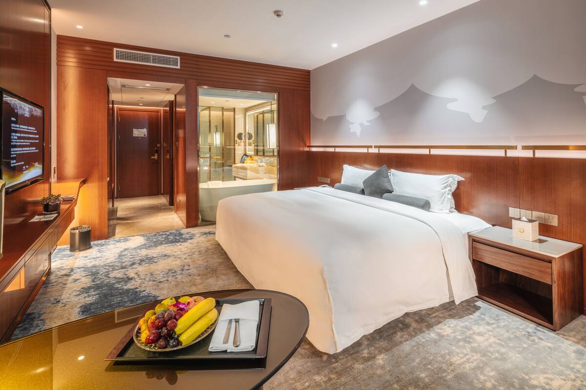 A hotel room with a large bed, a bedside table with a lamp, a TV, and a fruit tray on a small table.