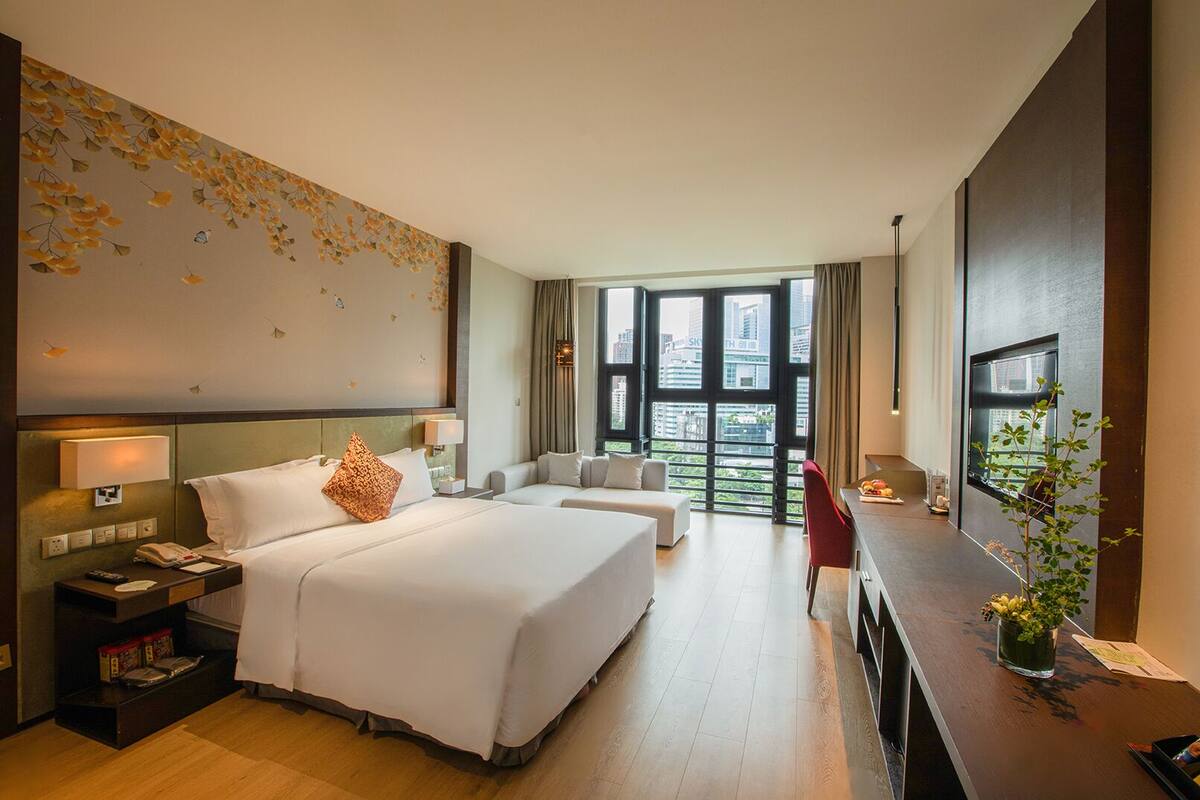 A hotel room with a large bed, a desk, a chair, and a view of the city through large windows.