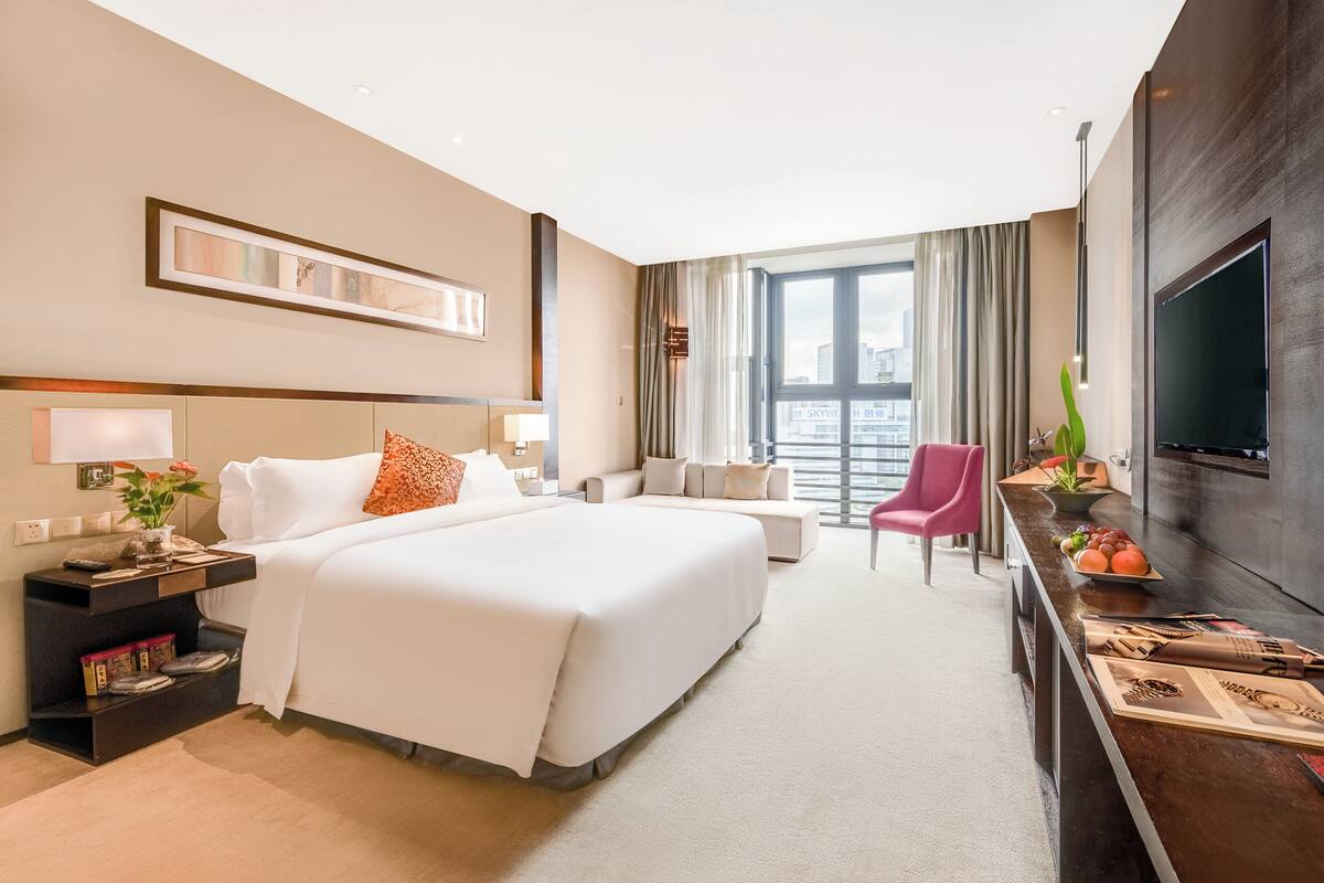 A hotel room with a large bed, a TV, a desk with a plant, and a view of the city.