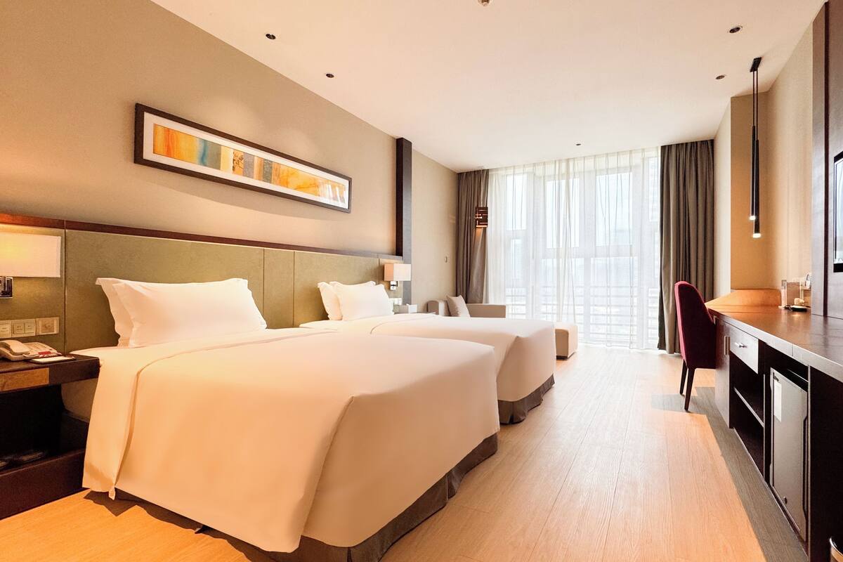 A hotel room with two beds, a desk, a chair, a TV, and a large window with curtains.