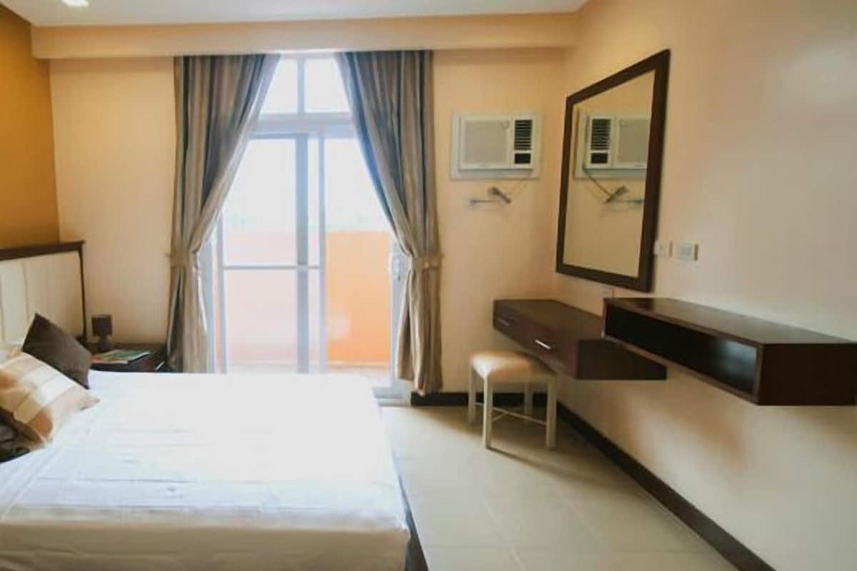A hotel room with a bed, a desk, a chair, a mirror, and an air conditioning unit.