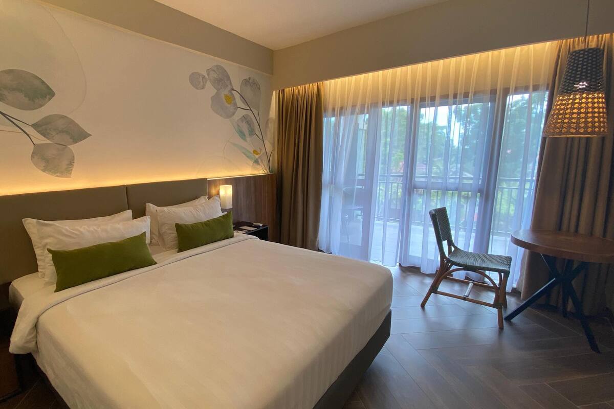 Ein Hotelzimmer mit einem großen Bett, einem Nachttisch, einer Lampe, einem Sessel und Blick ins Freie durch transparente Vorhänge.