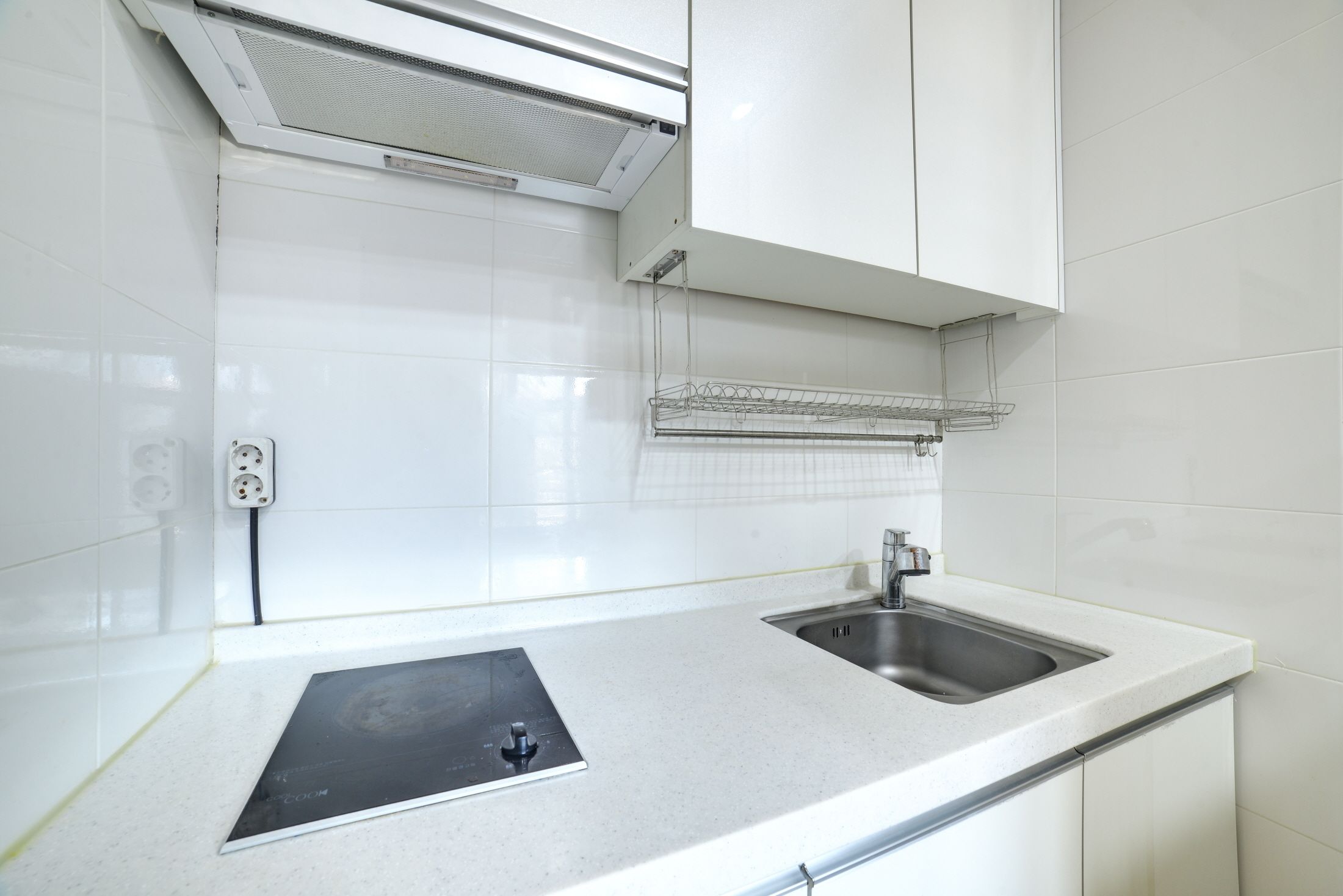 A kitchen with white tiles, a stainless steel sink, a black induction cooktop, and a ceiling-mounted ventilation system.