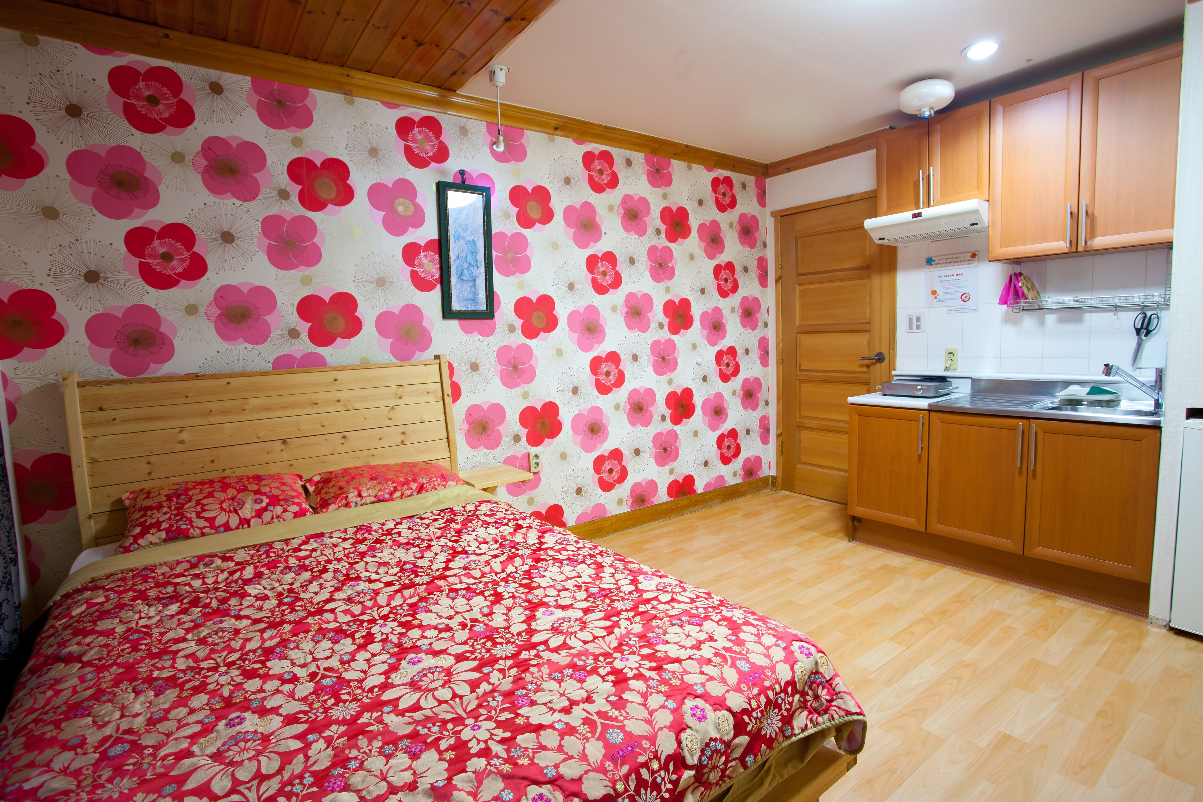 Un dormitorio con cabecera de madera, colcha con estampado floral y una zona de cocina con armarios de madera y un refrigerador blanco.