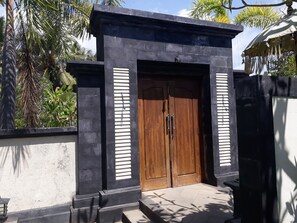 A wooden double door with a black frame, flanked by vertical black panels, leading to a building with a white exterior wall. - Pandawa Village (Sukasada)
