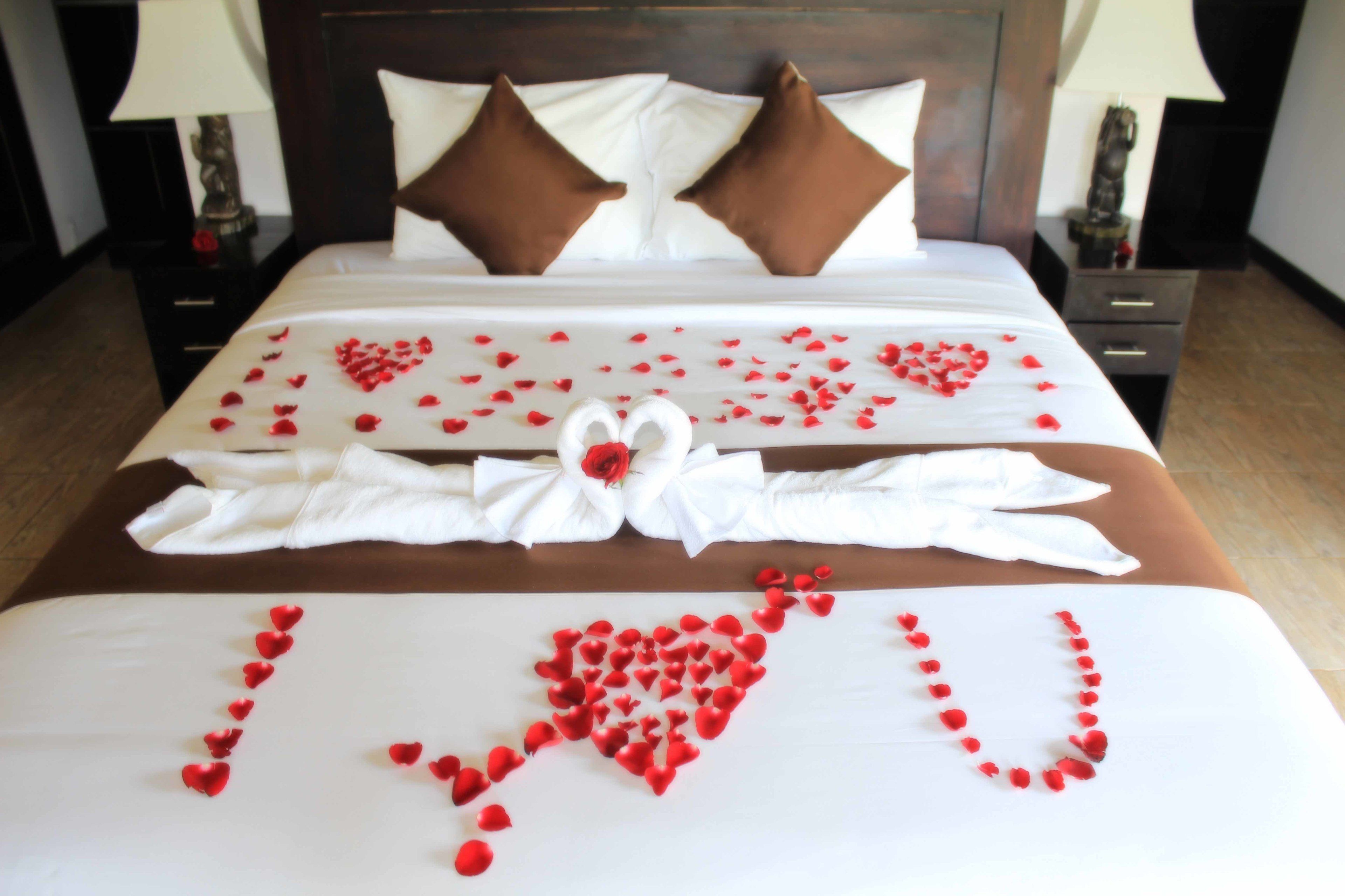 Una cama impecable con pétalos de rosa dispuestos en forma de corazón que deletrea “Te amo” y una toalla blanca con un lazo.