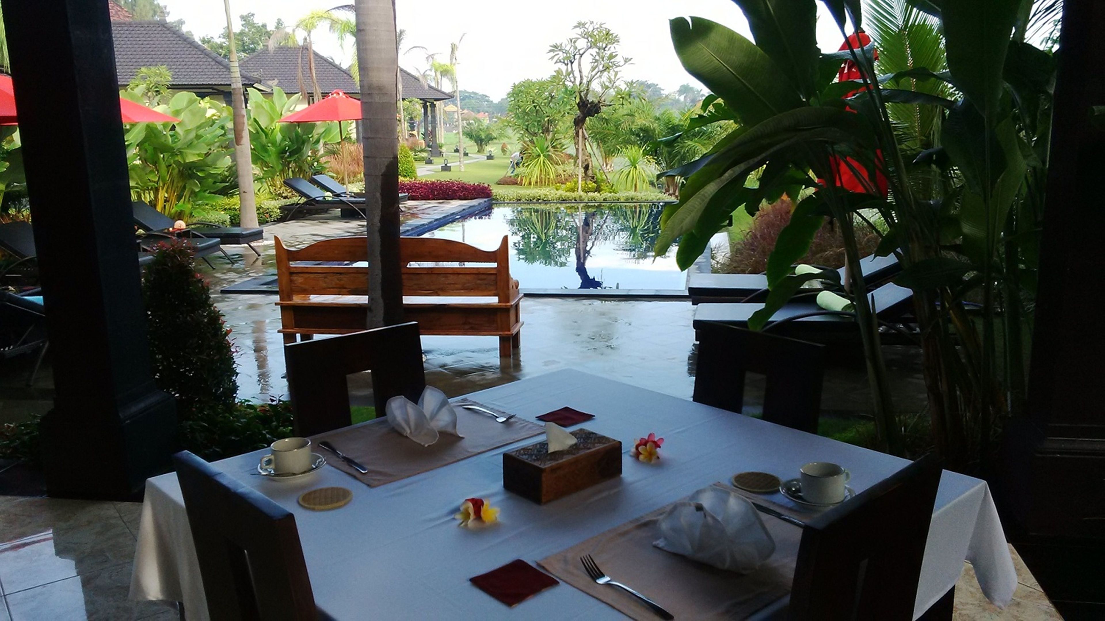 A dining area with a table set for two, overlooking a pool and lush greenery.