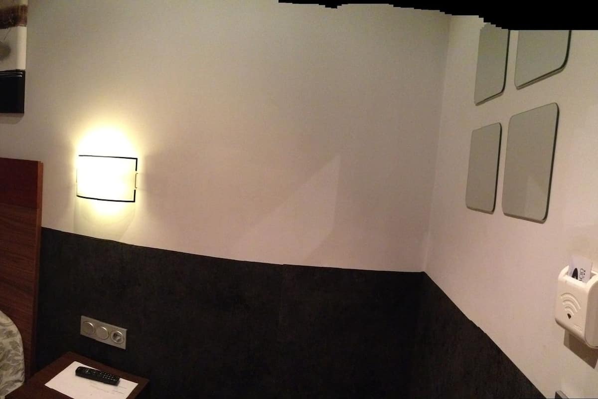 A hotel room with a bed, a bedside table, a chair, a small table, a mirror, a window with curtains, and a wall-mounted light fixture.