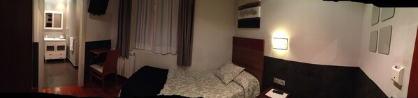 A hotel room with a bed, a bedside table, a chair, a small table, a mirror, a window with curtains, and a wall-mounted light fixture.