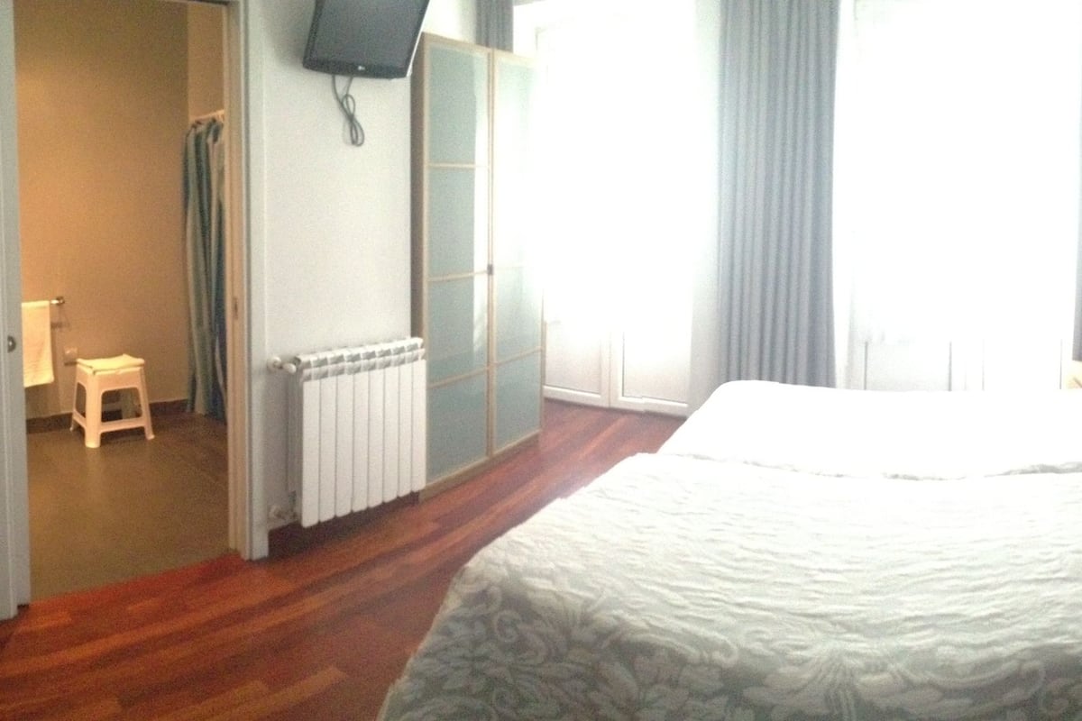 A hotel room with a bed, a chair, a desk, a radiator, and a television on the wall.