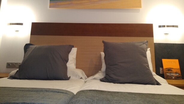 Une chambre d’hôtel avec un lit agrémenté de deux oreillers, une table de chevet sur laquelle repose un livre, et un tableau représentant une scène de plage, placé au-dessus du lit.