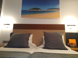 A hotel room with a bed featuring two pillows, a nightstand with a book, and a painting of a beach scene above the bed. - Pensión San Sebastián Centro (San Sebastián)