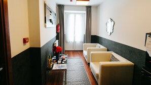 Une chambre d’hôtel avec un canapé, une table basse avec des magazines, une horloge au mur et une fenêtre avec des rideaux.