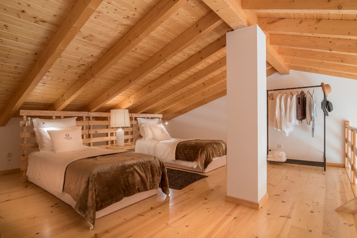 Un dormitorio con dos camas, vigas de madera en el techo, una pared blanca y un perchero con ropa colgada.