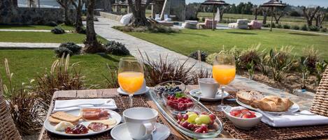 An outdoor breakfast setting with a table set with various breakfast items, including toast, eggs, and fruits, overlooking a garden and pool area.
