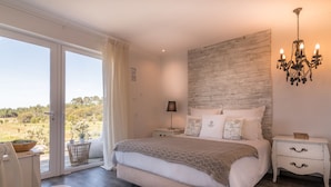A bedroom with a large bed, a chandelier, a nightstand, a chair, and a view through a glass door. - Monte do Cardal (Aljezur)