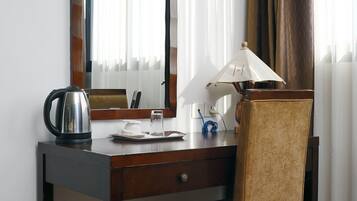 A hotel room with a wooden desk, a chair, a mirror, a lamp, and a kettle.