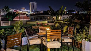Ein Sitzbereich im Freien mit Holzkästen und -tischen, umgeben von Blumentöpfen und Blick auf die nächtliche Skyline.