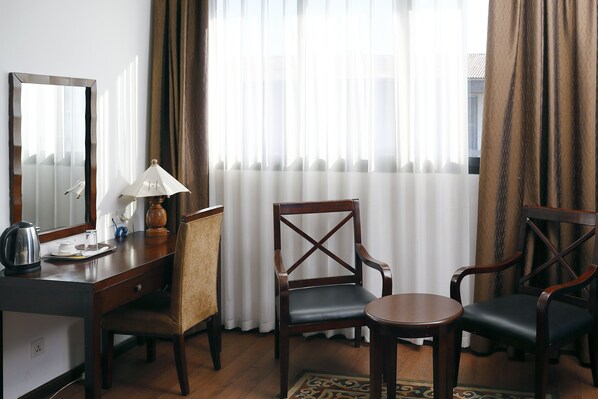 Habitación de hotel con un escritorio de madera, dos sillas, una lámpara, un espejo y vistas a través de cortinas translúcidas.
