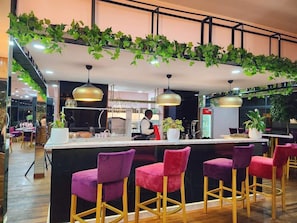 Zona de bar con mostrador, taburetes morados, plantas colgantes e iluminación colgante.