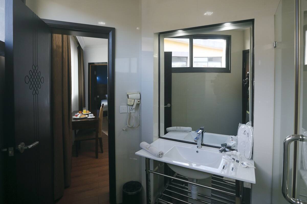 Une salle de bain d’hôtel avec un grand miroir, un lavabo et des toilettes. On aperçoit, au fond, une petite table avec une chaise et un téléphone.
