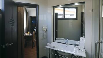 A hotel bathroom with a large mirror, a sink, and a toilet. There is a small table with a chair and a phone in the background.