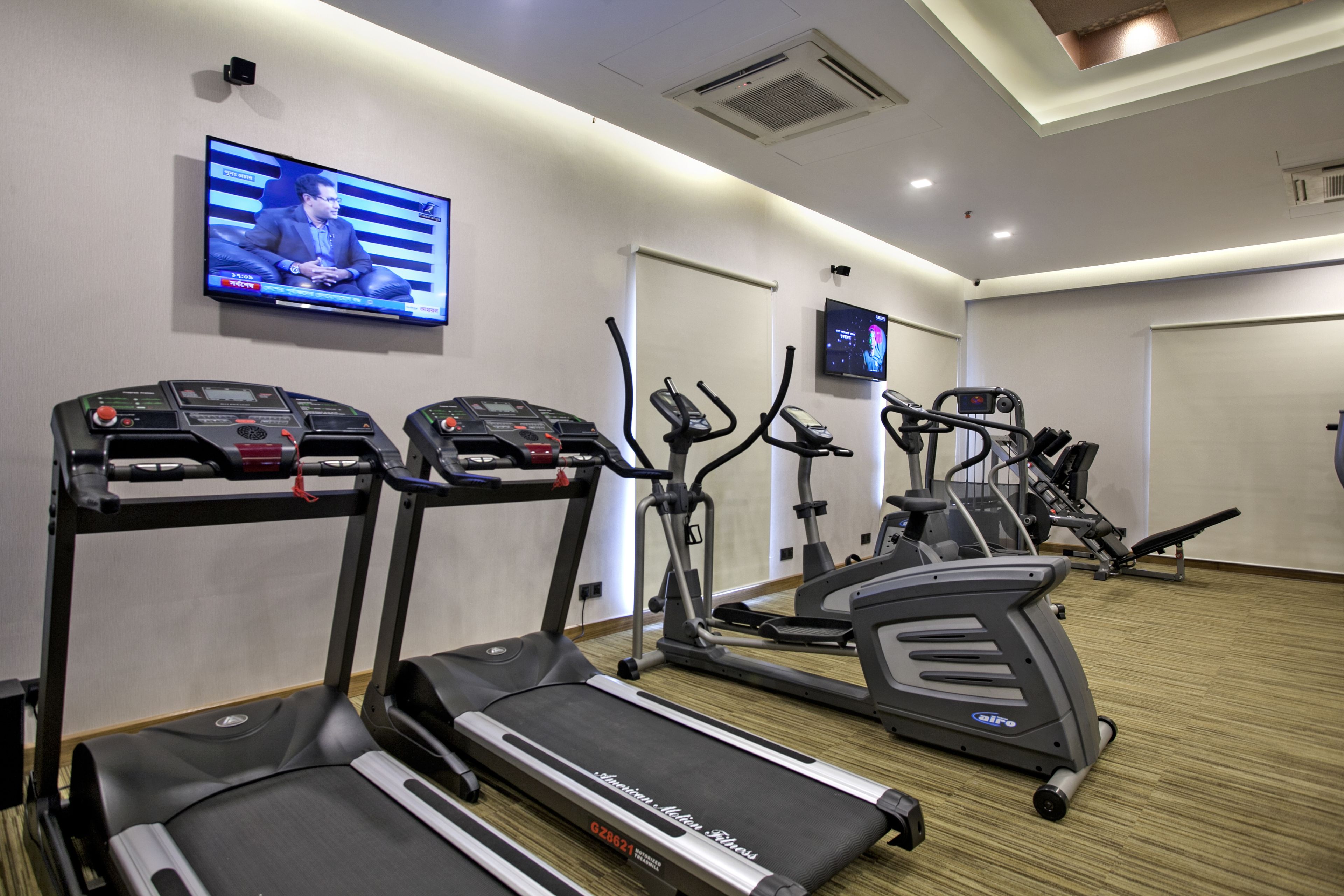 A gym with treadmills, ellipticals, and a TV displaying a news program.