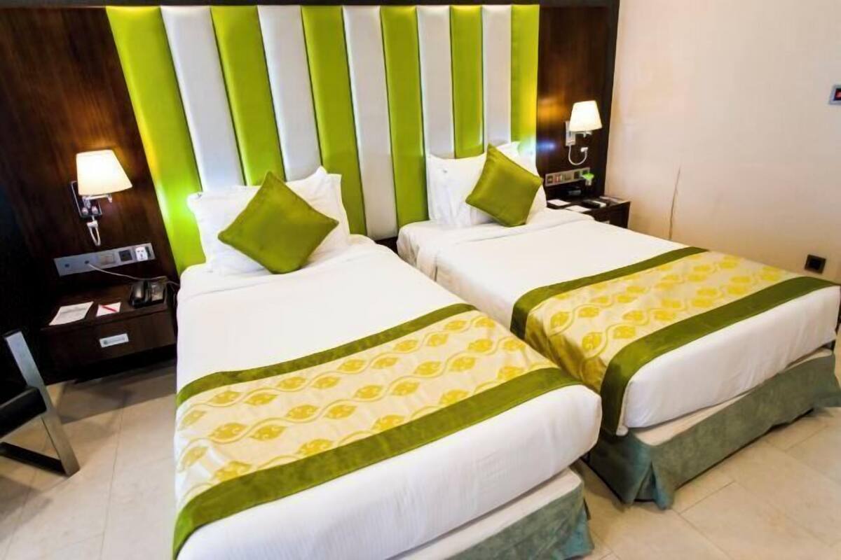Une chambre d’hôtel avec deux lits, chacun recouvert d’une couvre-lit verte et blanche, dotée d’une tête de lit à rayures, de lampes de chevet et d’une table de chevet sur laquelle se trouvent un téléphone et des documents.