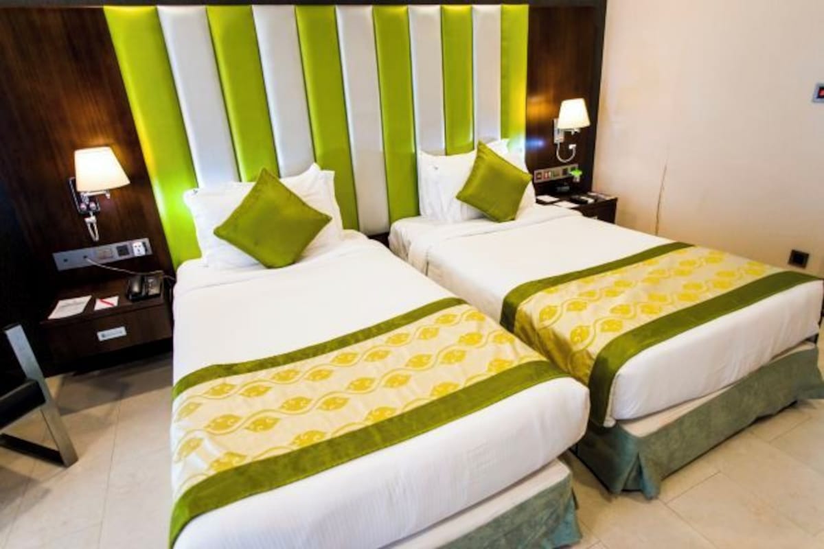 A hotel room with two beds, each with a green and white bedspread, a striped headboard, bedside lamps, and a nightstand with a phone and papers.