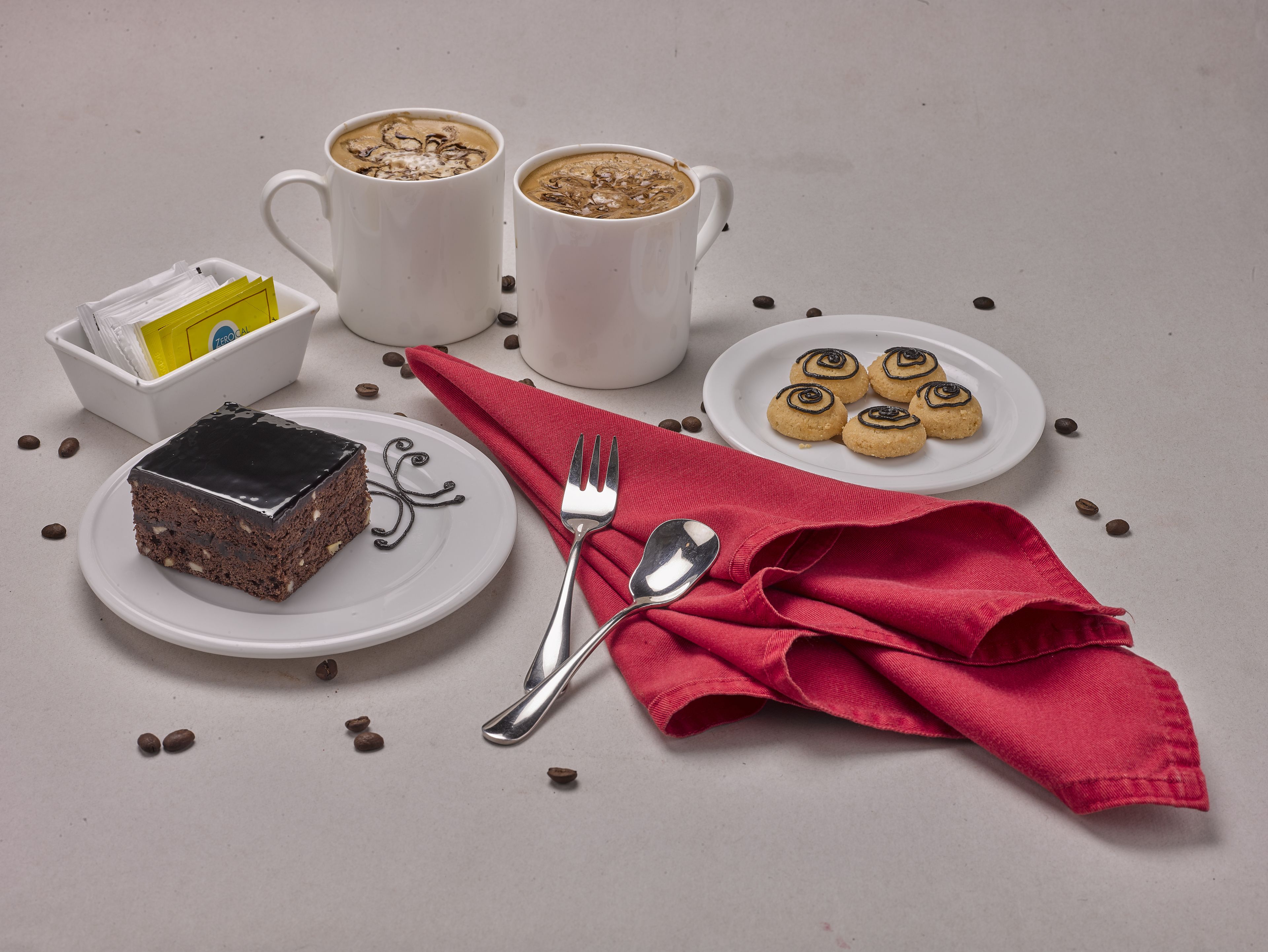 A table with a slice of chocolate cake, two cups of coffee, a plate with cookies, and scattered coffee beans.