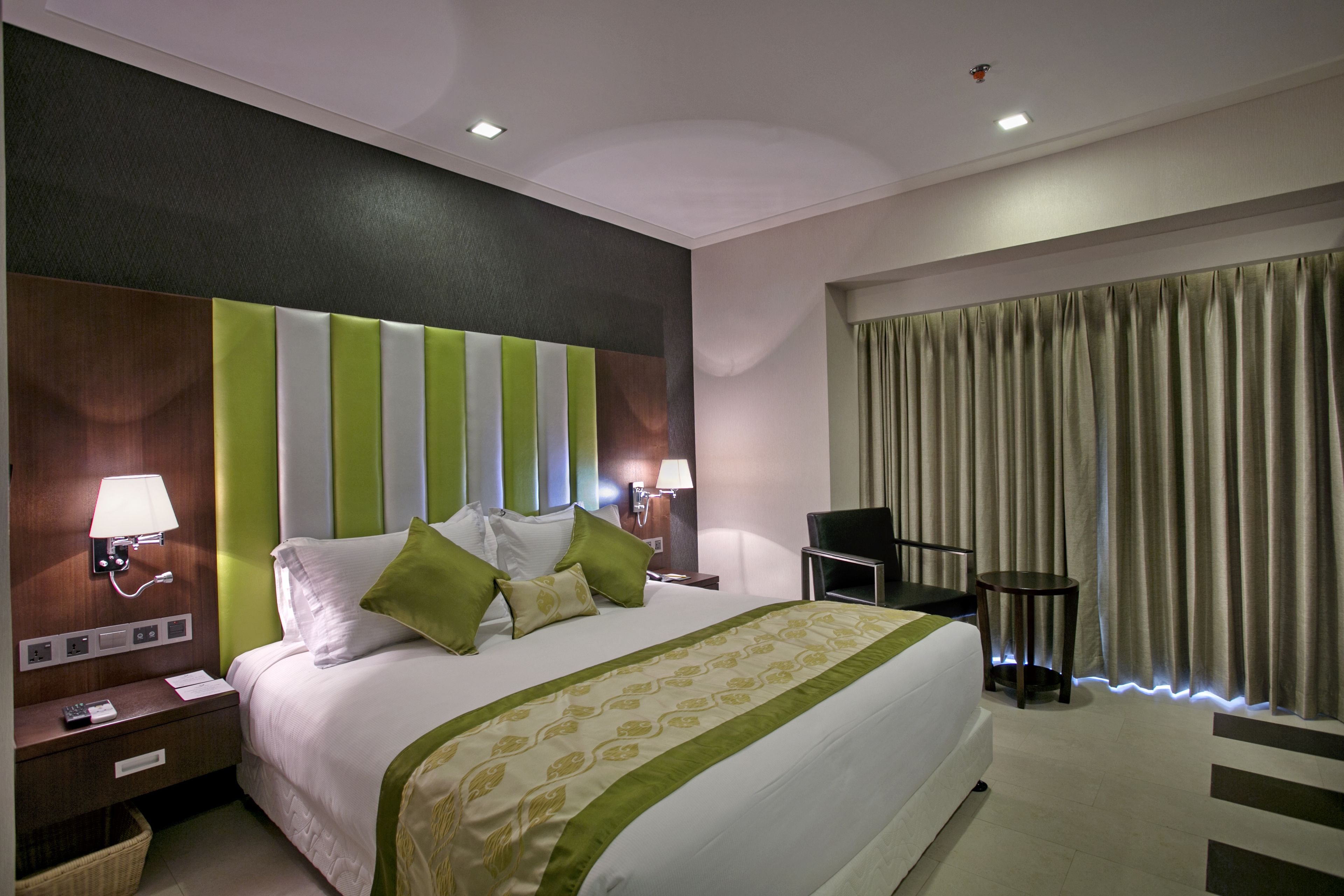 A hotel room with a large bed, green and white striped headboard, two bedside lamps, a small round table, and a window with curtains.
