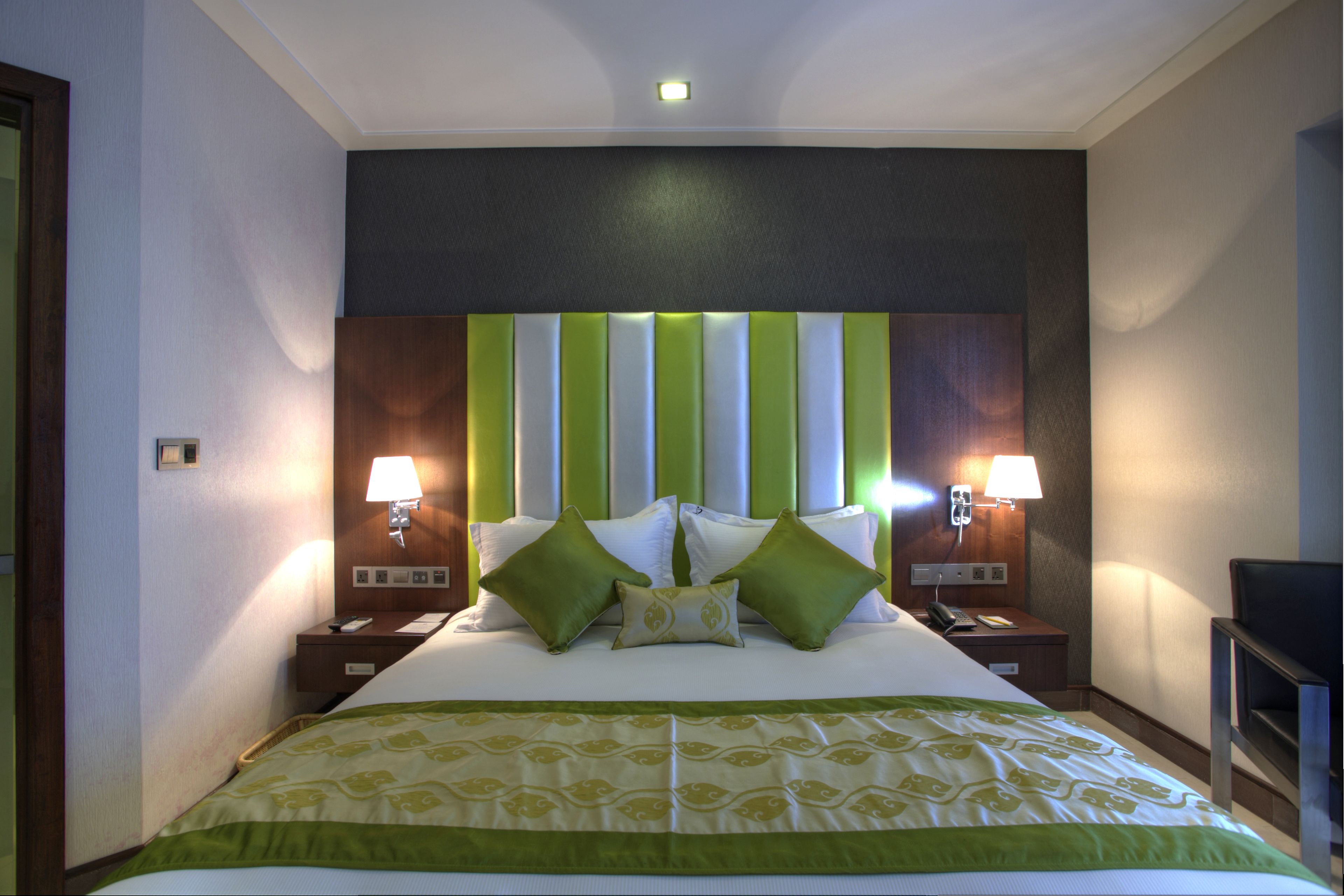 Habitación de hotel con una cama grande, cabecera verde y blanca a rayas, dos lámparas de noche y un pequeño escritorio con un teléfono.
